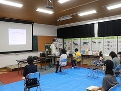 写真：青いビニールシート上に間隔をあけて椅子に座って話を聞いている会場の様子