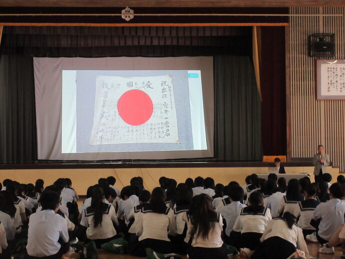 スクリーンに映し出された日本国旗を見つめる磐田北高校の生徒たちの様子の写真